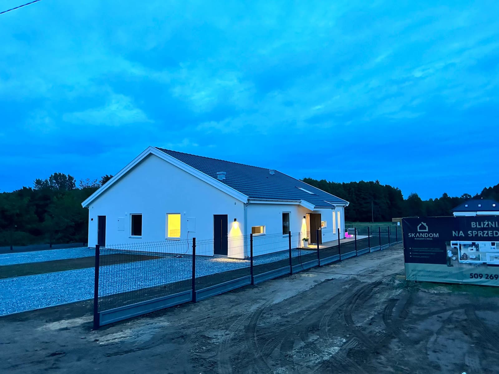 Dom o zmierzchu — rozświetlone okna i ogrodzenie od strony drogi