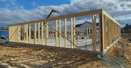 Drewniany szkielet ścian zewnętrznych ustawiony na płycie fundamentowej, w tle plac budowy